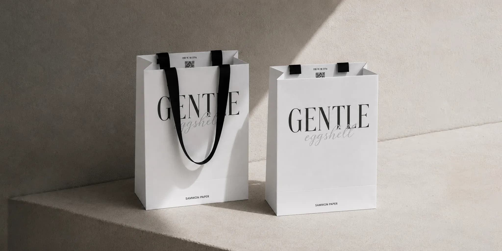 Two white paper bags with "GENTLE eggshell" printed on them, featuring black handles, placed on a concrete surface in soft lighting.