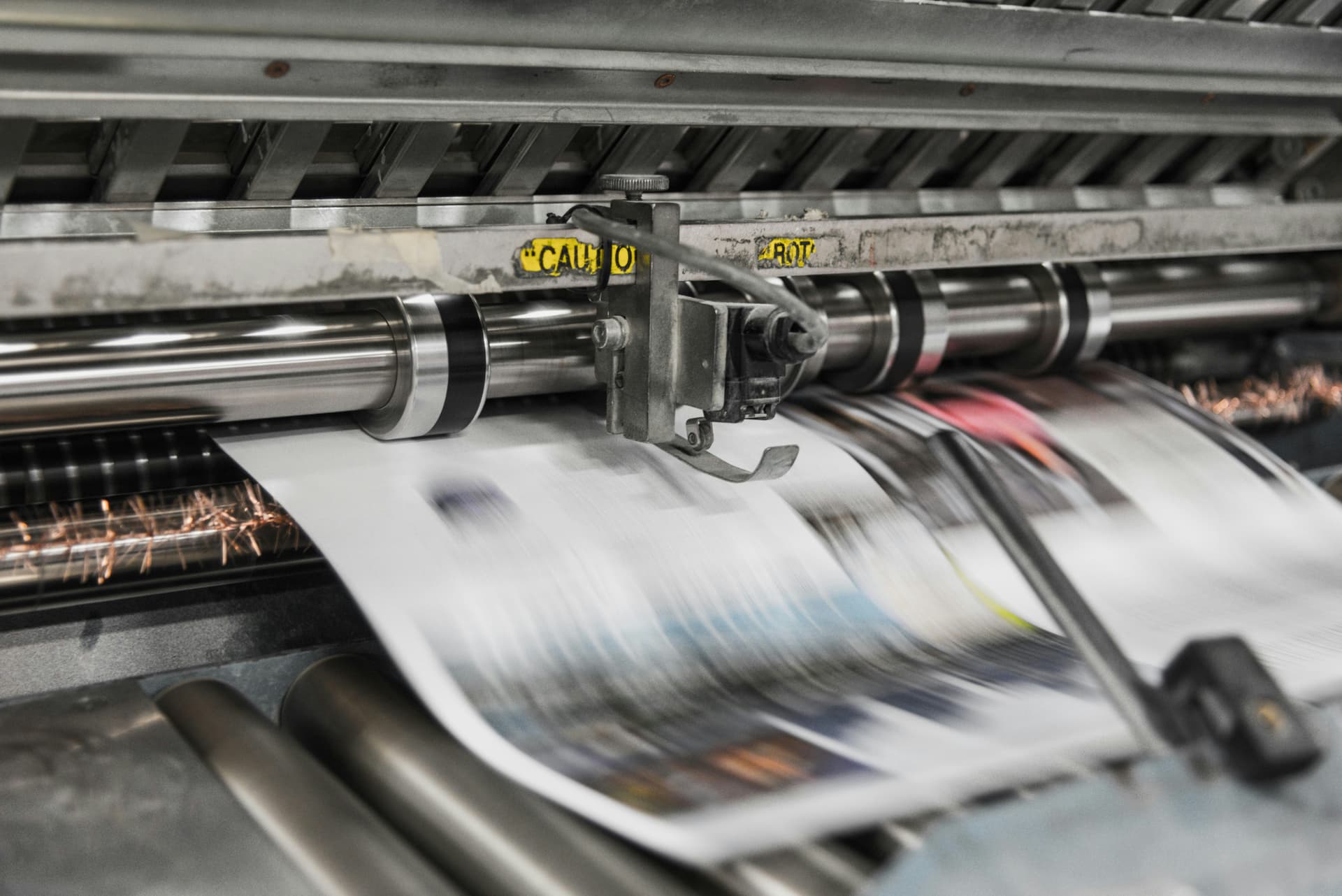 Close-up of a printing press in motion, rapidly producing color-printed pages with visible caution labels on machinery.