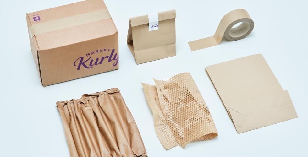 Various eco-friendly packaging items, including a cardboard box, paper bag, tape, crumpled paper, cushioning wrap, and an envelope, all in neutral tones.